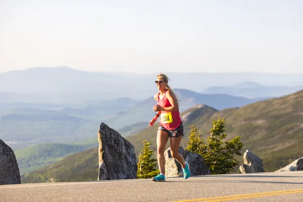 A female runner wearing a tank top, shorts, and sunglasses smiles while running during the Whiteface Mountain Uphill Race.