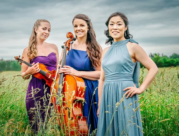 Three women in elegant dresses stand in a field. One holds a cello, another a violin.
