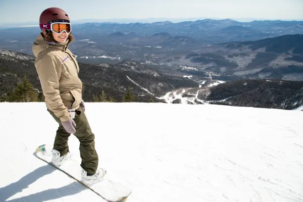 A smiling snowboarder at the top of a snowy run overlooking the main lodge of Whiteface Mountain.