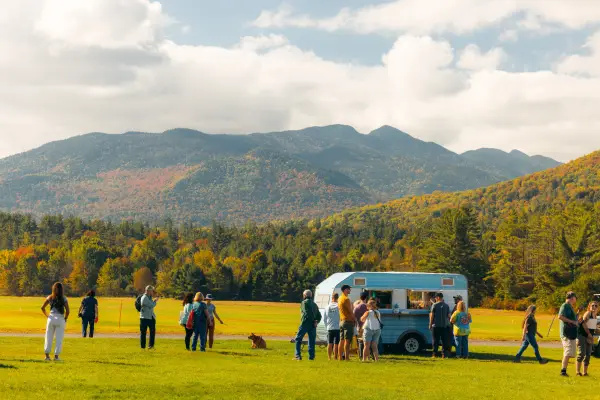 People stand around blue and white striped camper trailer in field with mountains in the background showing fall colors