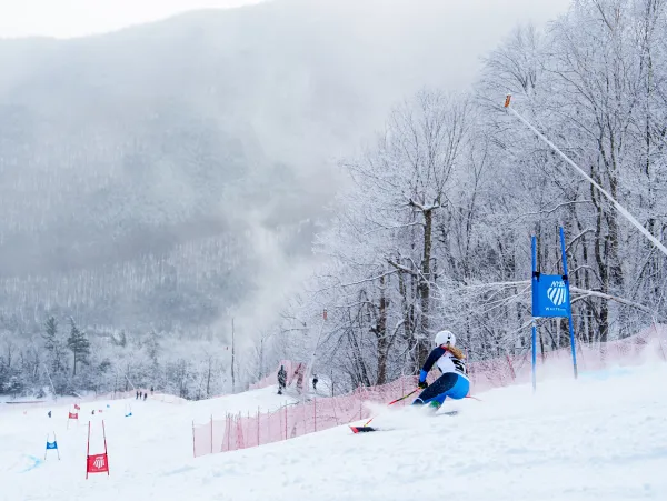 An alpine ski racer in a blue and black speed suit carves through a turn on a snow-covered mountain slope.