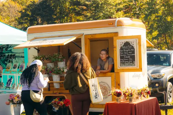 Two women browse flowers in metal buckets hung on side of camper trailer with woman leaning out window talking to customers