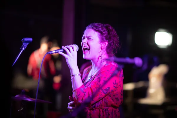 Woman in long sleeve red dress sings into microphone on dark stage