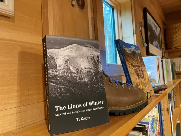 Book titled "The Lions of Winter: Survival and Sacrifice on Mount Washington" by Ty Gagne, shown on a wooden shelf with a hiking boot and other books.