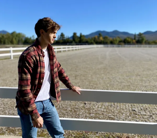A young man in a flannel and jeans stands next to a white fence.