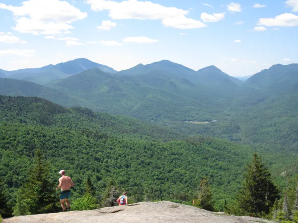 A shirtless man and a woman wearing a backpack on top of a rocky outcropping with the lush treed mountains surrounding