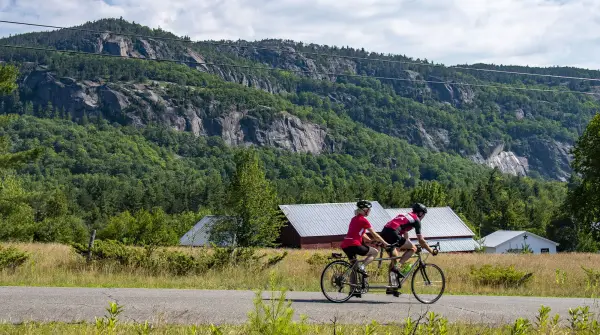 Tandem cyclists on road with barn and cliffs in background
