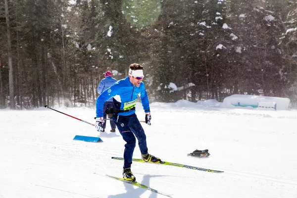 A skier in blue and black cross-country ski gear skis past a course worker during the Lake Placid Loppet race. The skier has a yellow race bib on their chest.
