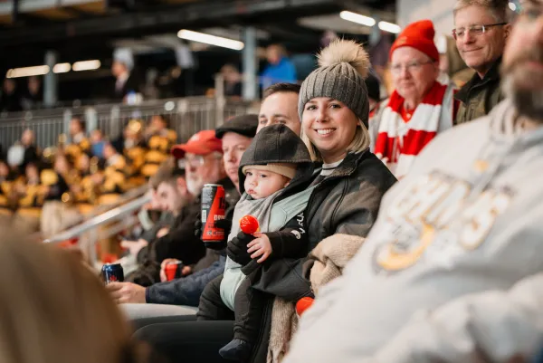 Woman sits amid crowd at hockey game in 1980 Herb Brooks Arena with baby in handsfree carrier on her chest. She smiles at the camera, holding a drink in one hand and wearing winter clothing.