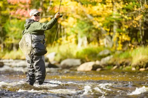 A man casts his line into the Au Sable River with bright fall foliage in the background