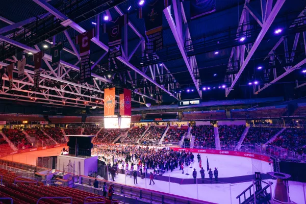 A wide-angle, high-angle shot captures a crowded ice arena during the 2025 Empire State Winter Games opening ceremony, featuring athletes gathered on the ice under purple stadium lighting and championship banners hanging from the rafters.