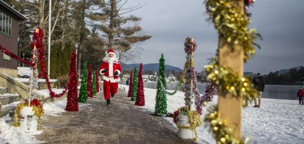A person in a Santa costume happily jogs along a snow-covered path lined with festive, colorful tinsel decorations and small tree figures.