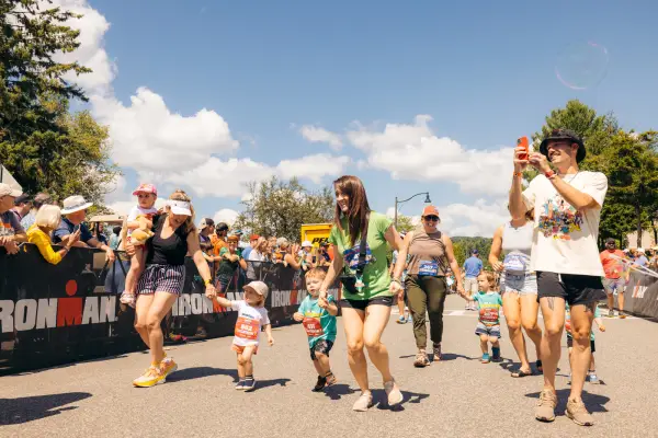 Collection of parents carrying some children and holding the hands of others walk on Ironkids race course between Ironman fencing lined with spectators