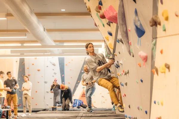 Climber ascends a bouldering route on an indoor bouldering wall while other climbers mingle in the background