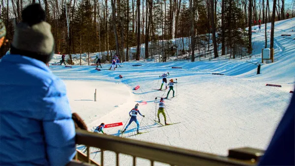 The image shows a winter sports scene with skiers racing on a snowy track surrounded by trees. 
