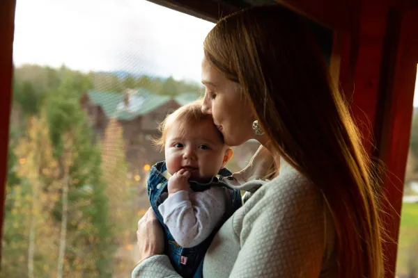 Woman kisses top of baby's head standing in large window overlooking trees and lodge-style resort