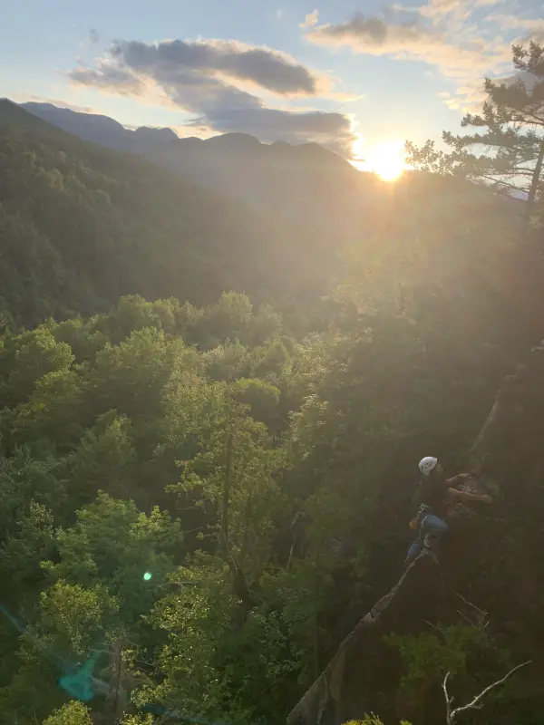 A rock climber ascends a cliff face silhouetted against a bright sunset and lush green forest.