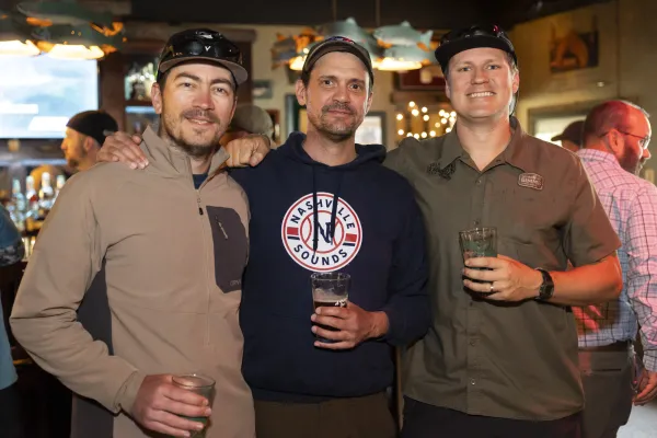 Three men stand together posing for photo with beer pints in hand at Ausable River Two-Fly Challenge event