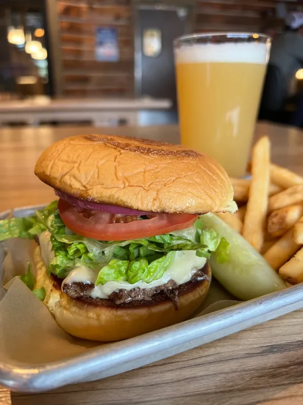 Cheese burger with thick slice of tomato and shredded lettuce with french fries, a pickle wedge and a freshly poured beer