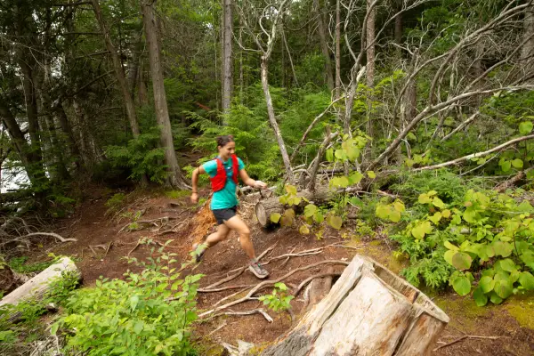 Woman in running shorts, running shoes, teal t-shirt, and red vest with hair in two buns runs uphill on forest trail covered in roots and rocks