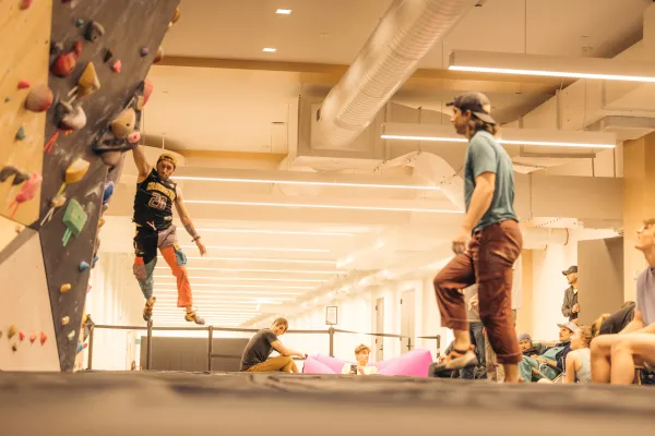 Climber hangs off of bouldering wall by one hand above black mats while other climbers look on