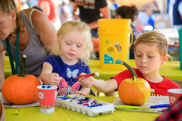 Young girl and young boy sit at table at event painting two small pumpkins