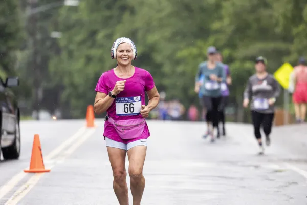 A smiling woman with light skin runs toward the camera during an outdoor race.