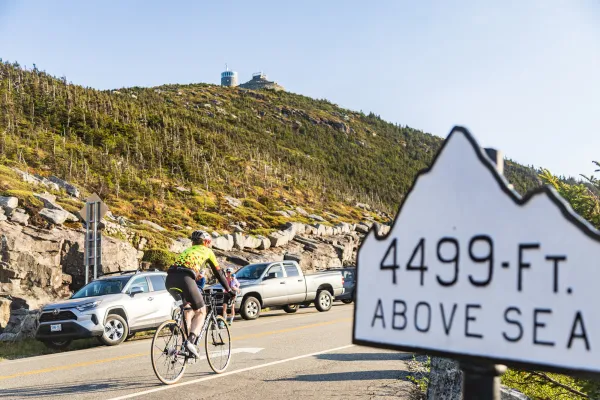 A cyclist rides uphill past parked cars on a mountain road, with a sign in the foreground reading "4499-FT. ABOVE SEA LEVEL".