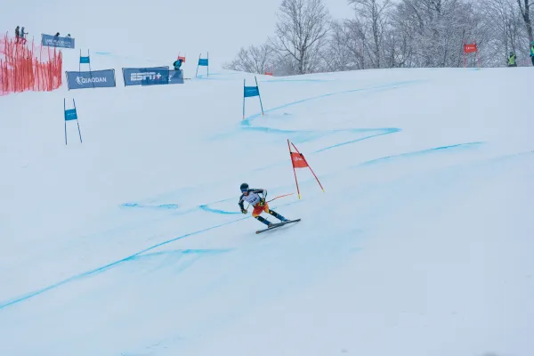 A downhill skier in a racing suit carves through a turn on a snowy slalom course marked by blue and red gates.
