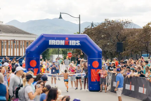 Boy in swim trunks runs toward finish line with blue inflatable arch while spectators cheer at Lake Placid Ironkids race