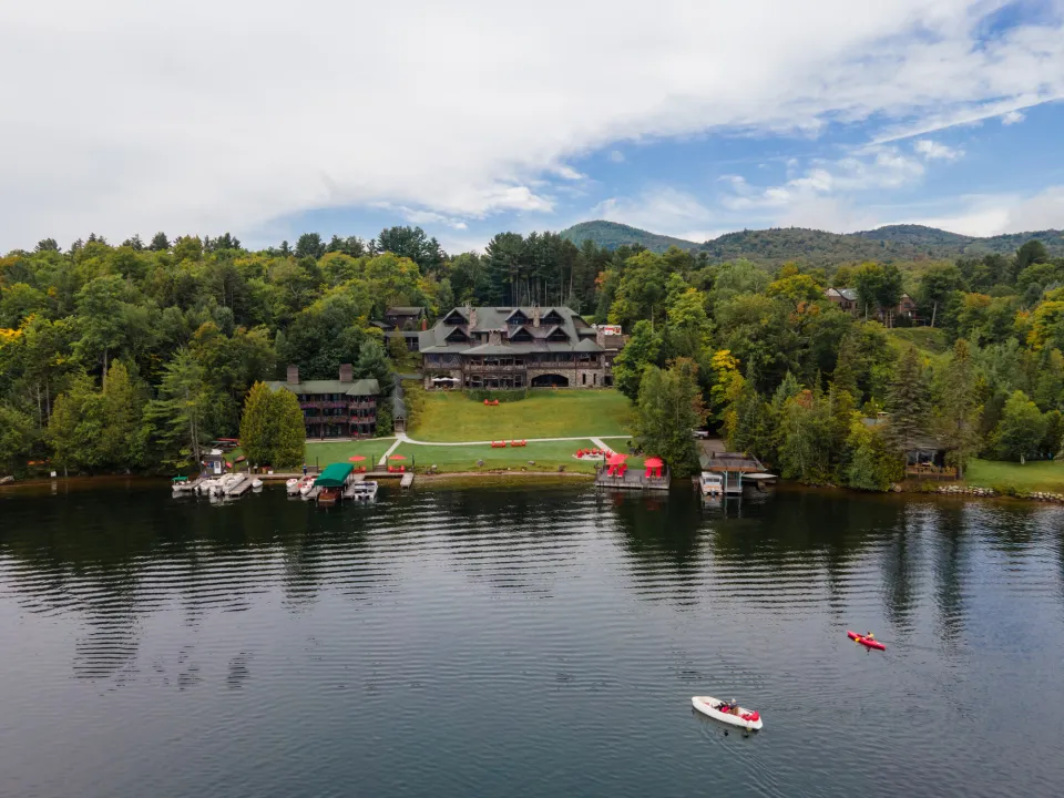 A grand Adirondack lodge sits at the edge of a lake. Two paths wind down the lawn to the water where boats sail. 