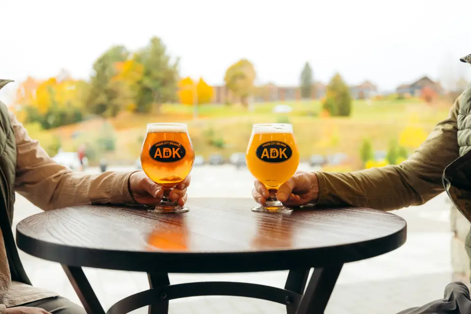 Two people holding beers on a table with fall scenery in the background.