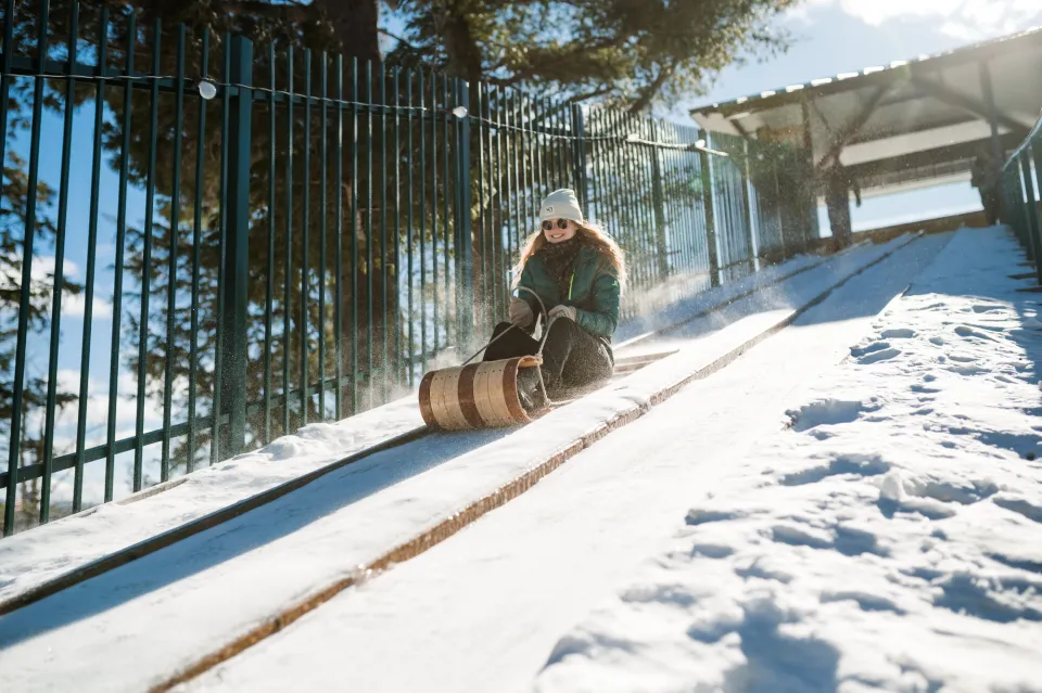 Woman in winter outdoor clothing slides down large manmade slide on toboggan