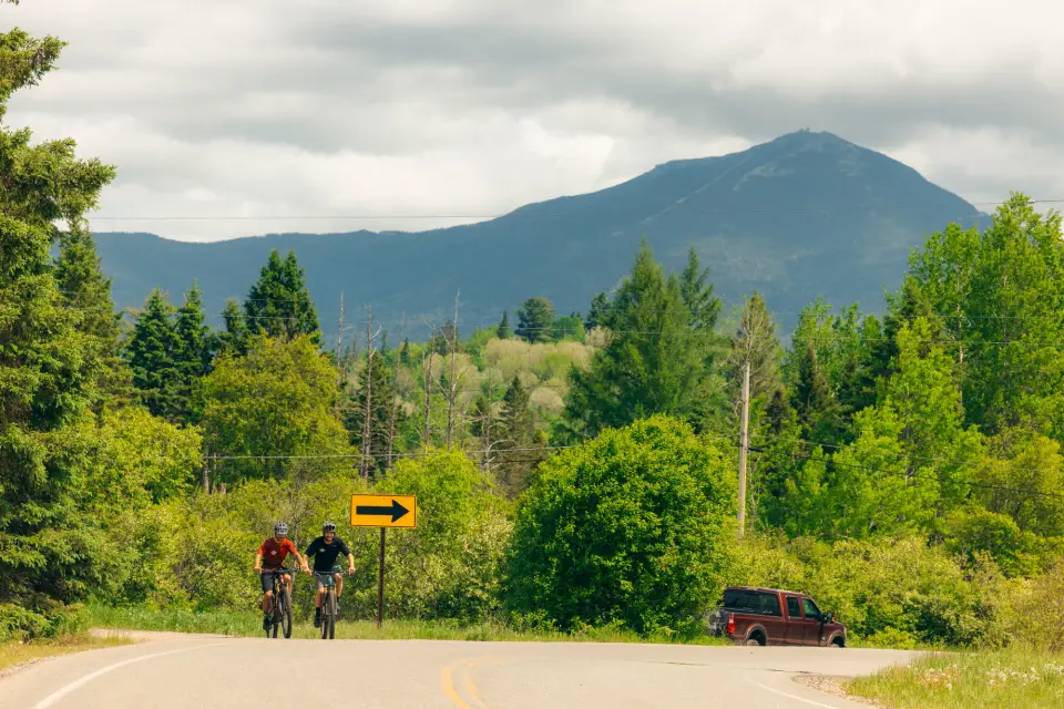 Two bikers ride side by side turning onto road toward camera while truck turns away from bikers driving away with forest and mountains in background