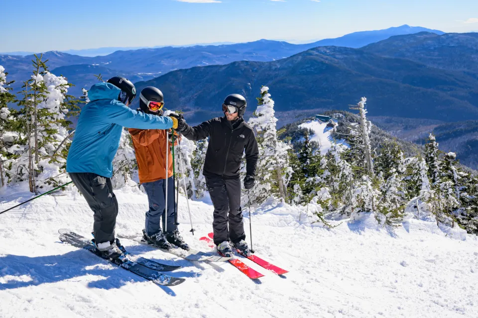 Three men fist bump in ski gear on top of a snowy mountain.