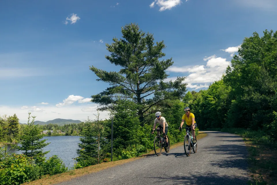 Two adults bicycle on a broad path alongside a strip of water, surrounded by pine trees.