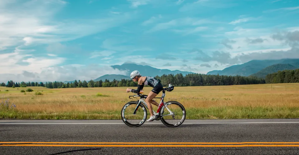 Ironman cyclist rides bike on rural road with field and mountains in background