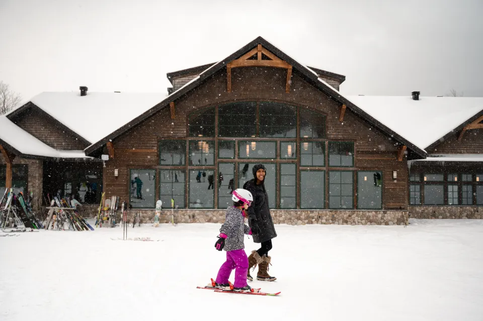 A woman walks her daughter on skis in front of a grand ski lodge in winter. 