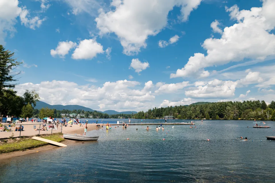 A wide view of a sparkling blue lake with a small sandy beach on the left. A dock extends into the water and swimmers are in the water. Low mountains are in the distance.