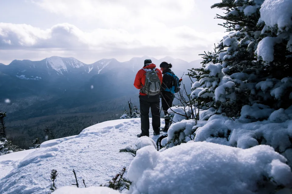 Two people stand at the top of a snowy summit while winter hiking in Lake Placid.