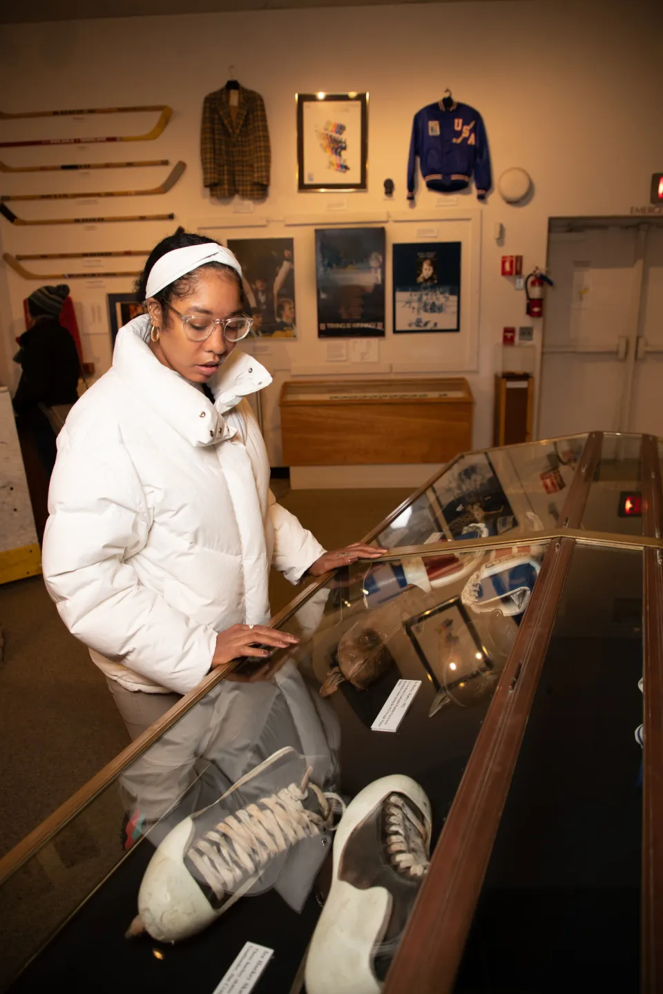 A woman in a white coat and glasses looks down at a glass case of Olympic memorabilia.