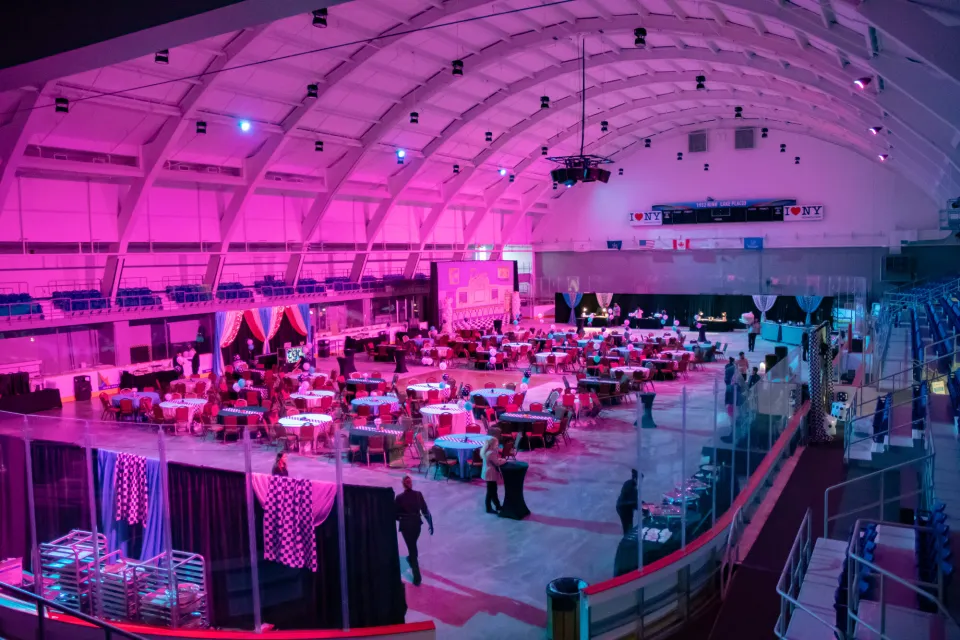 A white ice skating rink is lit up in pink light with white tables spread out for a dinner. 