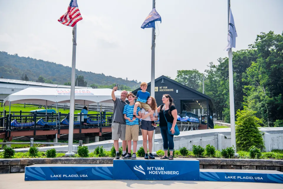 Family of five stands on first place block of podium holding youngest child aloft and taking a selfie