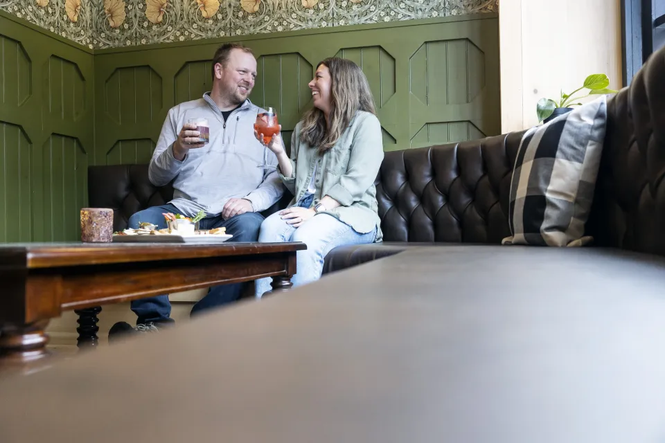 Couple sits closely in corner of bench seat with cocktails and a cheese board smiling at one another