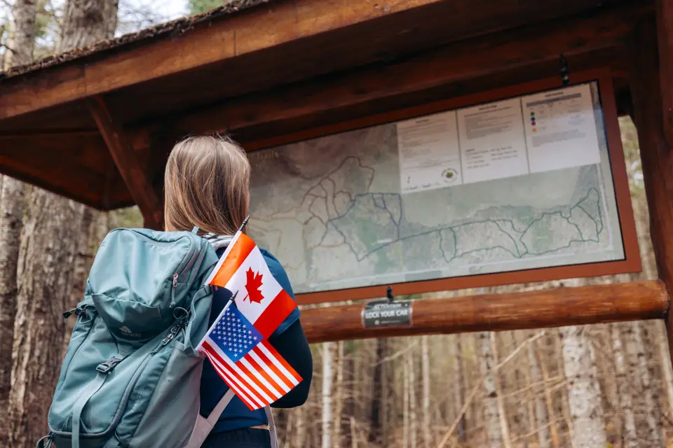A woman wearing a backpack looks at a hiking kiosk. Small USA and Canadian flags stick up out of the backpack.