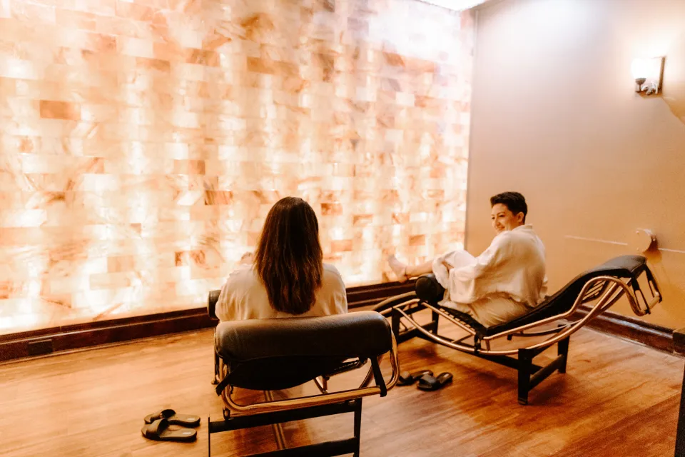 Two women lounge in spa loungers in front of a glowing salt wall in a luxury spa.
