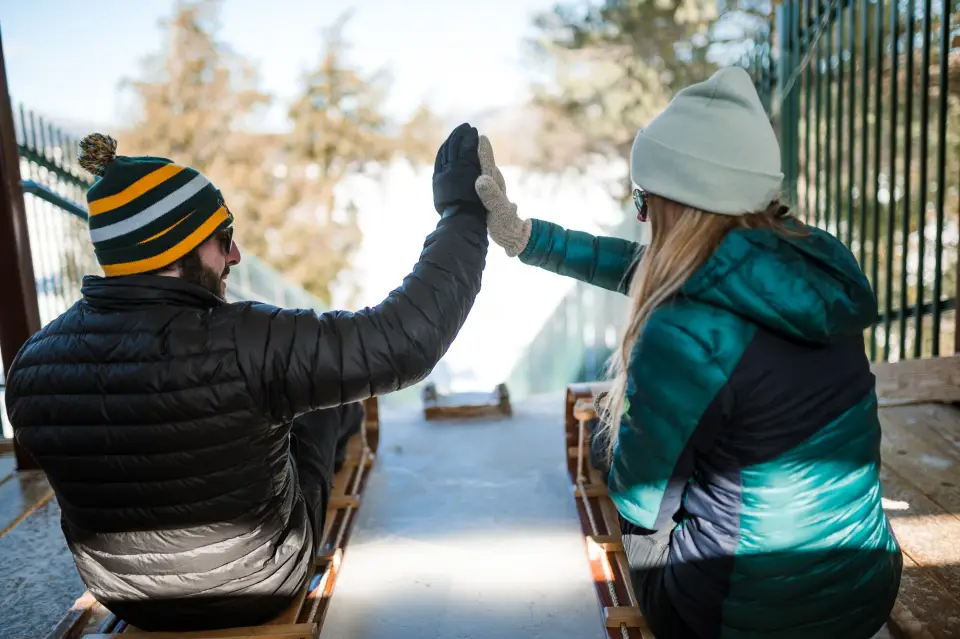 Two adults wearing winter clothing high five at the top of a toboggan chute.
