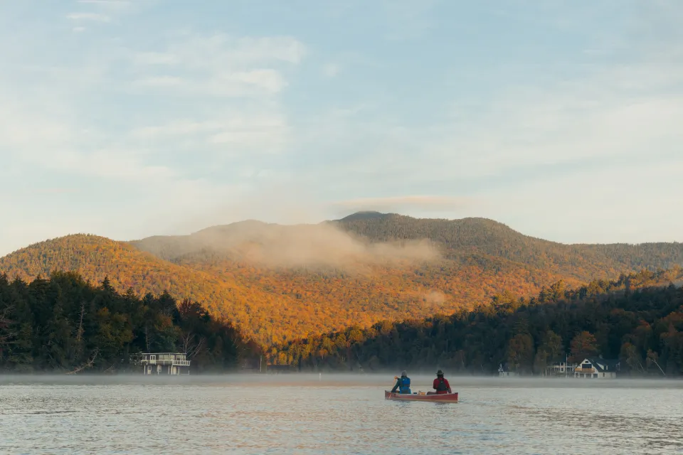 Two adults paddle a canoe on a calm lake at sunrise on a fall day.