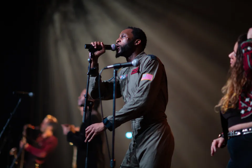 Man in green jumpsuit outfit stands on stage singing into microphone
