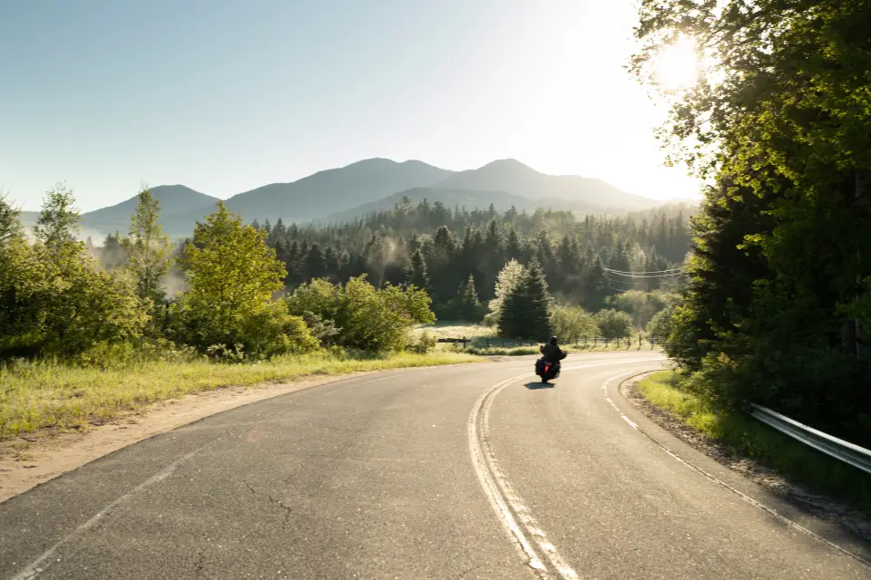 A single motorcycle leans into a curve in the road on a sunny day with forest and mountains in the background.